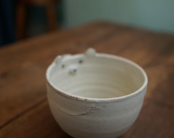 May include: A handmade, off-white ceramic bowl with a bear-like design on the rim. The bowl has a textured surface and a slightly irregular shape, suggesting a handcrafted origin. The bowl is set on a wooden surface.