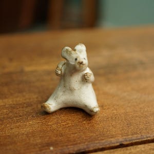 May include: A small, off-white ceramic figurine of a bear in a seated position. The bear has its arms raised and its ears are slightly pointed. The figurine is on a wooden surface.