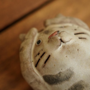 Może przedstawiać: Ceramiczna figurka kota w kremowym kolorze z szarymi plamami. Kot ma czarne wąsy, mały czerwony nos i zamknięte oczy. Figurka leży, z lekko pochyloną głową.