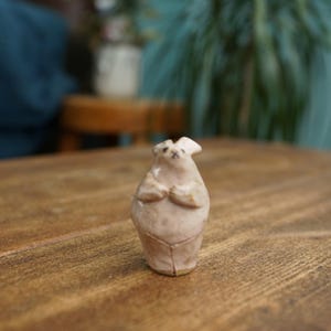 May include: A small, light-colored ceramic figurine of a bear with its paws clasped together. The bear has a simple, rounded form with small ears and a slightly textured surface. It is standing on a wooden table.