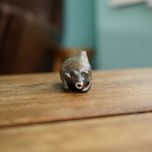 May include: A small, handcrafted bear figurine in a seated position. The bear is a dark brown color with a white nose and is positioned on a wooden surface. The figurine is a decorative item.