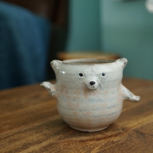 May include: A small, off-white ceramic mug shaped like a bear with two small ears, two outstretched arms, and a painted face featuring two black eyes and a black nose. The mug has a speckled, textured finish with subtle orange and blue undertones.