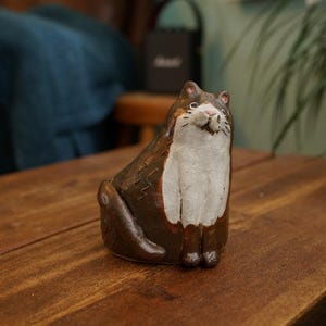 Può includere: Statuetta di gatto in ceramica marrone e bianca. Il gatto è seduto con le zampe unite e un'espressione sorridente. La statuetta è su una superficie di legno.