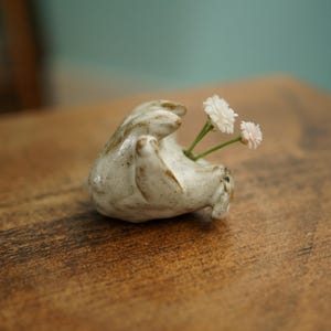 Peut inclure: Petit vase en céramique blanc cassé en forme de forme enroulée, contenant deux brins de fleurs blanches. Le vase a une surface texturée et repose sur une table en bois. L'arrière-plan est de couleur vert d'eau.