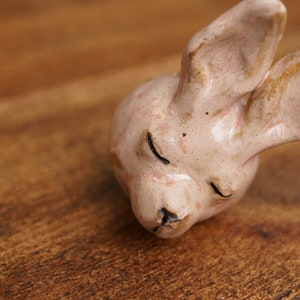 Op de afbeelding: Een keramieken konijnenkopfiguur in een zachte roze tint met zwarte ogen en een kleine zwarte neus. De oren van het konijn hebben een vleugje goud. Het beeldje rust op een houten oppervlak.