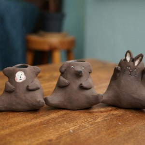 May include: Three brown ceramic animal-shaped planters. The first is a figure with a face, the second is a dog, and the third is a rabbit. Each has a small opening at the top for plants. They are sitting on a wooden surface.