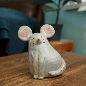 May include: A ceramic mouse figurine in a seated position. The mouse is light gray with pink accents on the ears. It has small black eyes and whiskers. The figurine is on a wooden surface.
