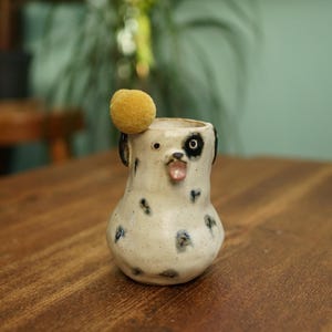 May include: A ceramic dog-shaped vase with a cream-colored base and black spots. The vase has a black and white eye, black ears, and a pink tongue. A small, yellow pom-pom sits on top of the vase. The vase is on a wooden surface.