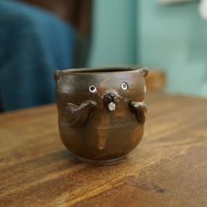 Könnte beinhalten: Ein brauner Keramikbecher in Form eines Bibers mit zwei kleinen weißen Augen, einer schwarzen Nase und einem einzelnen weißen Zahn, der aus der Mitte hervorsteht. Der Becher hat kleine geformte Ohren und zwei Vorderpfoten nahe dem Gesicht.