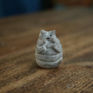 Op de afbeelding: Een klein, gebroken wit keramieken beeldje in de vorm van een gestileerde kat met een rond, gesegmenteerd lichaam. Het figuurtje heeft puntige oren, kleine armpjes bij de borst en eenvoudige donkere markeringen voor de ogen en de mond.