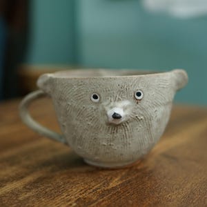 May include: A light gray ceramic mug shaped like an animal face with small ears, two round eyes, and a pointed nose. The surface features a textured, etched pattern throughout, and a single handle is attached to the side of the rounded cup body.