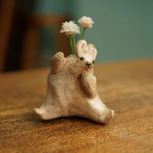 May include: A whimsical, off-white ceramic bunny-shaped vase holding two small white flowers. The bunny is in a seated position with its arms raised, and has a small black cross for a nose. The vase is on a wooden surface.
