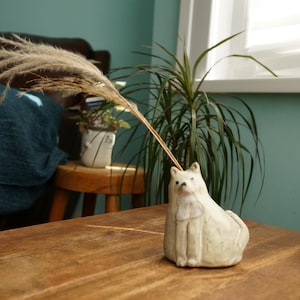 Puede incluir: Un jarrón de cerámica con forma de oso polar de color crema, que sostiene un tallo de planta seca. El jarrón está sobre una mesa de madera. Una manta azul oscuro y una planta de interior están en el fondo.