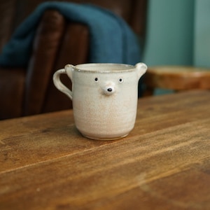 May include: A beige ceramic mug featuring a sculpted bear face with two small ears on the rim, two dark eyes, and a protruding nose. The mug has a single handle on the left side and a speckled, textured finish across the entire exterior surface.