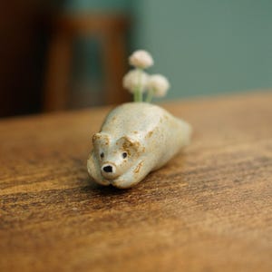 Puede incluir: Un jarrón de cerámica con forma de oso en color beige claro, con pequeñas flores blancas que emergen de su espalda. El oso tiene ojos negros y un hocico detallado. El jarrón está colocado sobre una superficie de madera.