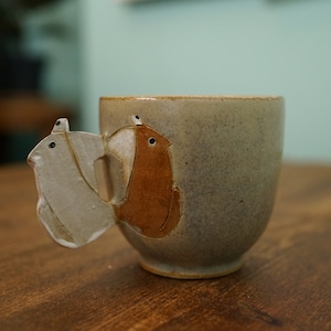 Może przedstawiać: Ceramiczny kubek z unikalnym uchwytem przedstawiającym dwie ptasie postacie. Kubek jest w kolorze nakrapianego beżu z brązowo-białym wzorem ptaka. Kubek stoi na drewnianej powierzchni.