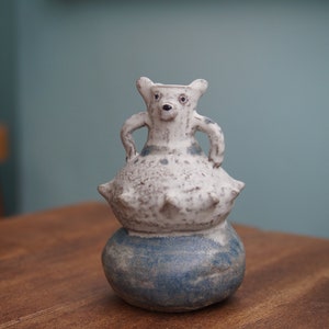 Puede incluir: Un jarrón artesanal de cerámica con forma de oso. El jarrón presenta un cuerpo moteado blanco y gris con una base azul. El oso tiene una pequeña nariz y orejas negras. El jarrón está sobre una mesa de madera.