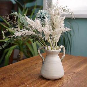 Puede incluir: Un jarrón de cerámica blanca con dos asas contiene un ramo de flores secas y vegetación. El jarrón está sobre una mesa de madera. El arreglo incluye hierba de la pampa blanca esponjosa, eucalipto y una flor azul.