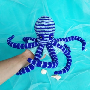 Op de afbeelding: Een handgemaakte gehaakte octopus in blauw en wit. De octopus heeft een gestreepte kop en tentakels, met een lichtblauwe franje. De ogen zijn zwart. Het is een zacht speelgoed.