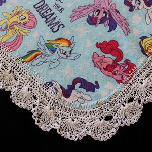 May include: A light blue blanket featuring cartoon ponies with a white crocheted lace trim. The blanket has the text "Chase Your Dreams" in white. The ponies are in various colors, including pink, yellow, and purple.