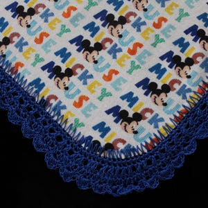 May include: A white fabric with a repeating pattern of Mickey Mouse faces and the word "MICKEY" in various colors. The edge is trimmed with a blue crocheted border.