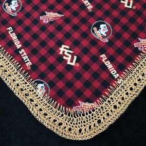 May include: A red and black plaid fabric with the words "FLORIDA STATE" and the Seminoles logo. The fabric is trimmed with a gold crochet lace border. The overall theme is collegiate.