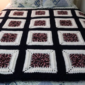 May include: A handmade crochet blanket in a square pattern. The blanket features alternating squares of navy blue and white, with a central cluster of burgundy and black yarn in each white square. The blanket is spread over a bed.