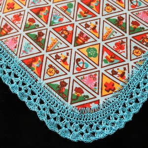 May include: A square fabric with a repeating pattern of colorful cartoon animals and shapes within triangles. The edge is trimmed with a turquoise crocheted lace border. The fabric has a light blue background.
