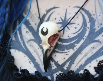 May include: A bird skull pendant necklace. The skull is off-white with a black beak and a crescent moon detail. The necklace is worn over a black top with a lace trim and a chest tattoo. The style is gothic.