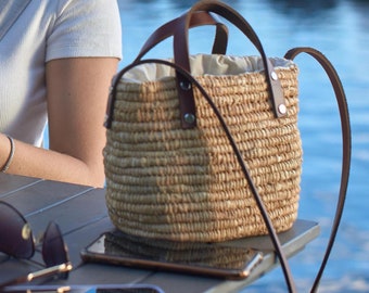 May include: A straw tote bag with brown leather straps and a light brown lining. The bag is sitting on a table with two cell phones and a pair of sunglasses.