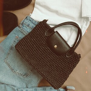 May include: A black woven handbag with a leather flap and a gold buckle. The bag is laying on top of blue jeans and a white t-shirt.