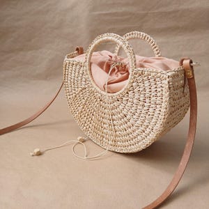 May include: A woven straw handbag with a rounded shape, featuring two circular handles and a long, brown leather strap. The interior is lined with a light pink fabric, and the bag has a drawstring closure. The bag is approximately 10 inches wide.