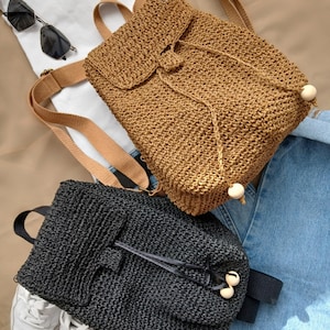 May include: Two woven straw backpacks with drawstrings and wooden toggles. One backpack is brown and the other is black.
