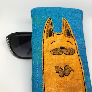 May include: A blue fabric eyeglasses case featuring a quirky embroidered orange cat with black eyes and a moustache. The cat is smiling and has a butterfly design on its chest.