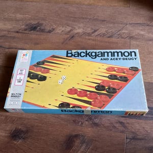 May include: A vintage Milton Bradley Backgammon game box with a yellow and black game board, red and black playing pieces, and two dice. The box has a red, white, and blue color scheme and the text "Backgammon and Acey-Deucy" printed on the front.