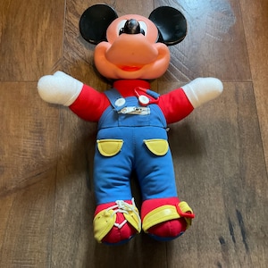 May include: A vintage Mickey Mouse plush doll. The doll has a peach-colored face, black ears, and a red shirt. It wears blue overalls with yellow accents and red shoes with yellow trim. The doll's arms are outstretched, and it has white gloves.