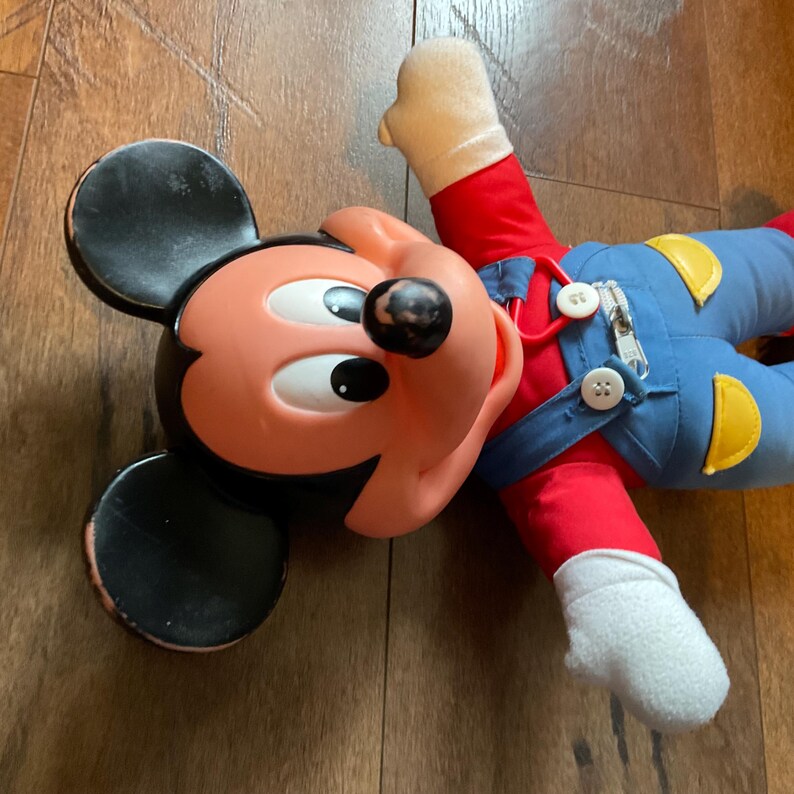 May include: A vintage Mickey Mouse plush doll. The doll has a black head and ears, a peach-colored face, and a red shirt. It wears blue overalls with yellow accents and white gloves. The doll is lying on a wooden surface.