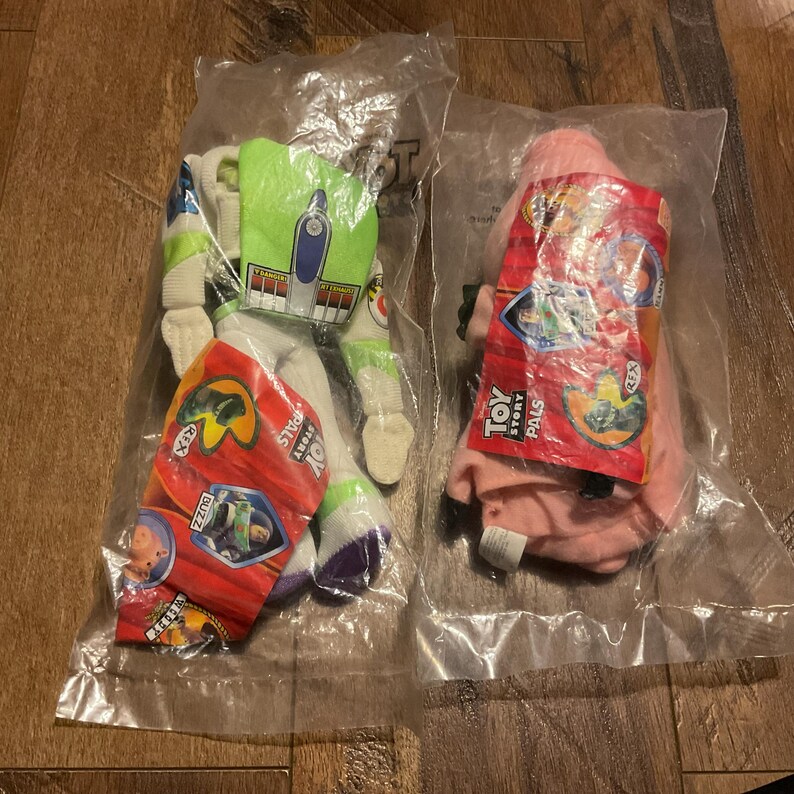 Może przedstawiać: Dwie pluszowe zabawki Toy Story Pals w oryginalnym opakowaniu. Pluszowa zabawka Buzz Lightyear jest zielona, biała i fioletowa. Pluszowa zabawka Rex jest r&oacute;żowa. Oba opakowania zawierają logo Toy Story Pals i postacie.