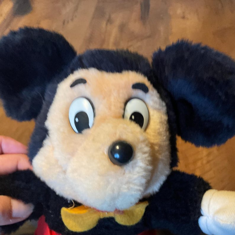 K&ouml;nnte beinhalten: Ein Pl&uuml;schtier von Micky Maus mit schwarzen Fellohren und -kopf, einem hellbraunen Gesicht und einer schwarzen Nase. Das Spielzeug hat eine gelbe Fliege und ein rotes Hemd. Das Spielzeug ist eine klassische Disney-Figur.