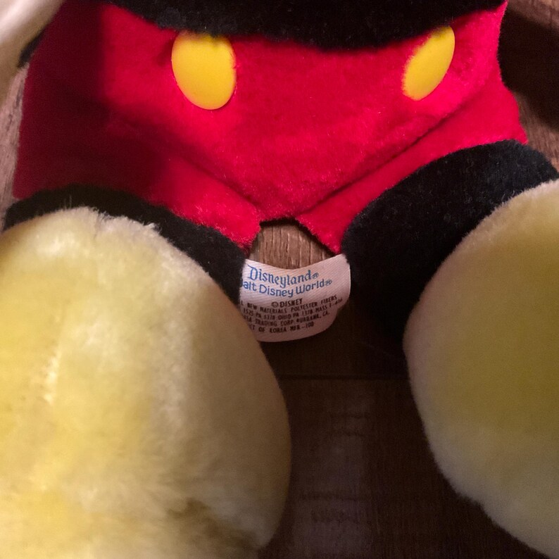 Pu&ograve; includere: Primo piano di un peluche vintage di Topolino. Il giocattolo presenta un corpo rosso con bottoni gialli, pantaloncini neri e grandi mani e piedi gialli. Un'etichetta recita "Disneyland Walt Disney World".