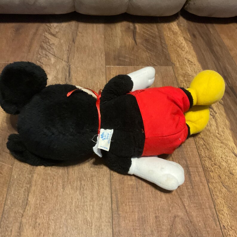 K&ouml;nnte beinhalten: Ein Stofftier von Mickey Mouse, das auf der Seite liegt. Das Spielzeug hat einen schwarzen K&ouml;rper, Ohren und Kopf, mit roten Shorts, wei&szlig;en Handschuhen und gelben Schuhen. Ein rotes Band ist um den Hals gebunden.