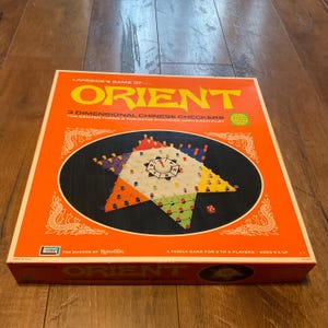 May include: A vintage board game box for "Orient," a 3-dimensional Chinese Checkers game. The box is orange with the game's name in large yellow letters. The game board features a star-shaped design with colorful pegs and a central clock-like element.