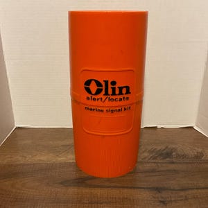 May include: Orange cylindrical container with black text that reads "Olin alert/locate marine signal kit".