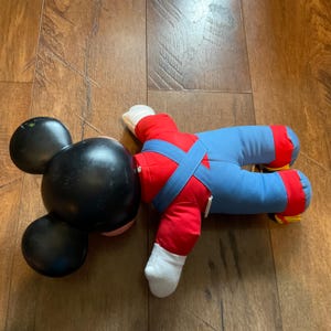 May include: A vintage Mickey Mouse plush toy, lying on a wooden surface. The toy features a large black head with round ears, red arms, blue overalls, and white gloves and feet. The plush is in a relaxed pose.
