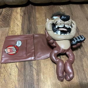 May include: A brown plush Tazmanian Devil toy with a brown leather seat cover. The seat cover has a pocket with the words "Taz Hungry!" printed on it. The toy is holding a remote control with buttons labeled "A, B, C, D, E, F, G, H, I, J, K, L, M, N, O, P, Q, R, S, T, U, V, W, X, Y, Z".