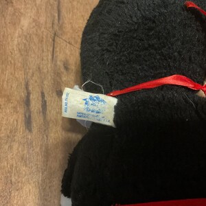 K&ouml;nnte beinhalten: Ein schwarzes Pl&uuml;schtier mit rotem Band und wei&szlig;em Etikett. Das Etikett hat blauen und wei&szlig;en Text. Das Spielzeug befindet sich auf einer Holzoberfl&auml;che. Auf dem Etikett steht "Hasbro, Inc. Pawtucket, R.I. U.S.A."