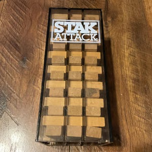 Peut inclure: Une bo&icirc;te en plastique transparent contenant un jeu de puzzle en bois. Le jeu est &eacute;tiquet&eacute; "STAK ATTACK" en texte blanc sur fond noir. Les pi&egrave;ces du puzzle sont rectangulaires et beiges.