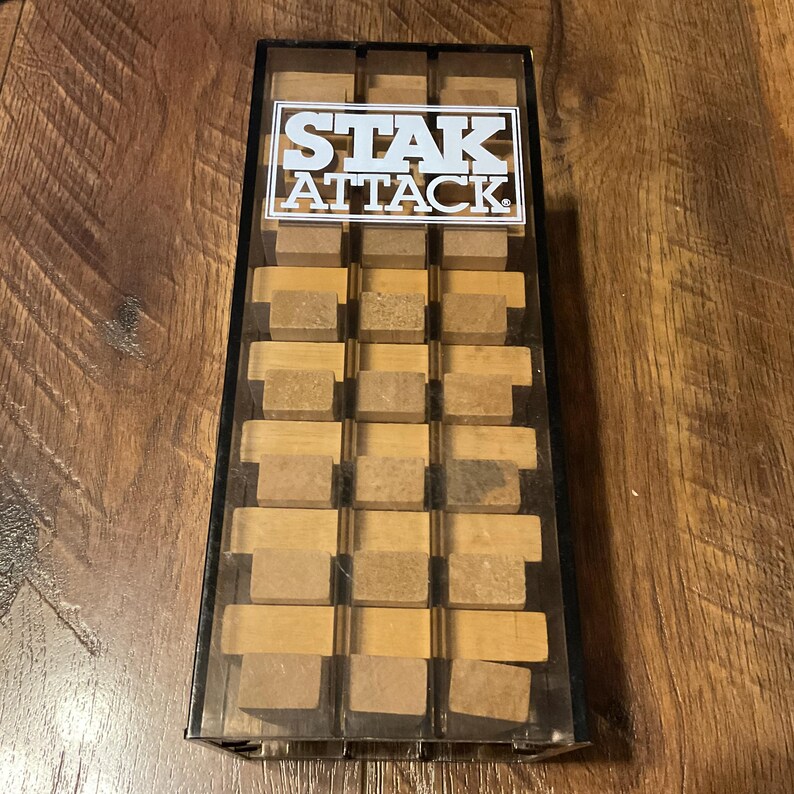 Peut inclure: Une bo&icirc;te en plastique transparent contenant un jeu de puzzle en bois. Le jeu est &eacute;tiquet&eacute; "STAK ATTACK" en texte blanc sur fond noir. Les pi&egrave;ces du puzzle sont rectangulaires et beiges.