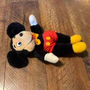 K&ouml;nnte beinhalten: Ein Pl&uuml;schtier von Mickey Mouse, das auf dem R&uuml;cken liegt. Mickey ist schwarz mit einem beigen Gesicht, roten Shorts mit gelben Kn&ouml;pfen, gelben Schuhen, wei&szlig;en Handschuhen und einer gelben Fliege. Eine Hand ist mit einem wei&szlig;en Handschuh und einer blauen Manschette erhoben.