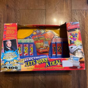 May include: A blue and yellow board game box with the title "Electronic Let's Make A Deal". The game features the voice of Monty Hall and includes a game board with a car, money, and numbers. The box also features a picture of a red sports car.